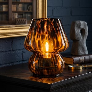 May include: A mushroom-shaped table lamp made of amber-colored glass with a dark, mottled tortoiseshell pattern. The lamp features a rounded base and a wide, flared shade, with a single glowing light bulb visible inside the glass structure.