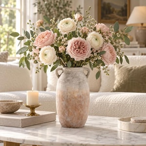 May include: A tall, two-handled ceramic vase with a textured, ombre finish transitioning from a light grey top to a warm, earthy terracotta base, filled with a lush arrangement of white and soft pink ranunculus flowers and green foliage.