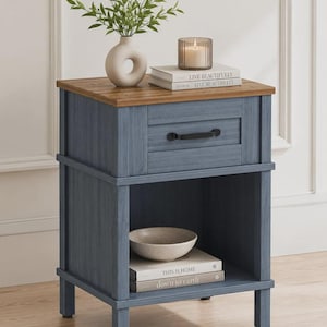 May include: A blue wooden nightstand with a natural wood-toned top, featuring one drawer with a black handle and an open lower shelf. The top holds a beige vase with greenery, a candle, and two books labeled Live Beautifully. A bowl and two books labeled This is Home and down to earth sit on the shelf.