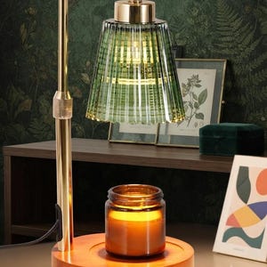 May include: A candle warmer lamp featuring a gold-toned metal stand, a ribbed green glass shade, and a circular wooden base. A brown glass jar candle sits on the base, illuminated by the warm light emitted from the lamp's overhead bulb.