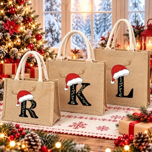 Può includere: Tre borse in juta color beige con manici color crema. Ogni borsa presenta una grande lettera nera, R, K o L, decorata con luci colorate e sormontata da un cappello da Babbo Natale rosso e bianco con corna di renna.