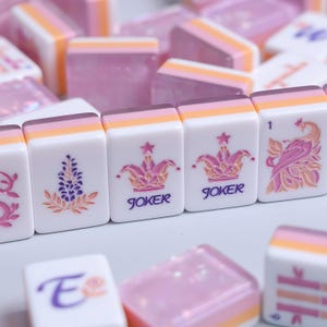 以下が含まれることがあります： 白を基調とし、ピンク、オレンジ、紫のアクセントが施された長方形の麻雀牌のセット。牌には、花の小枝、JOKERの文字が入った王冠、数字の1が刻まれた孔雀など、さまざまな彫刻デザインが施されています。