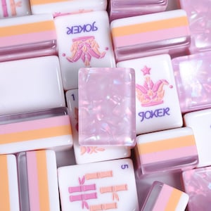Può includere: Una collezione di tessere da mahjong che include tessere bianche con strisce orizzontali rosa e arancioni, tessere con una grafica a corona rosa e la scritta JOKER, tessere con simboli di bambù rosa e una tessera centrale rosa traslucida.