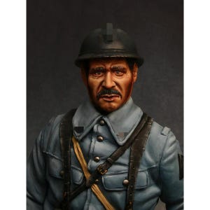 May include: A detailed bust of a soldier wearing a dark grey helmet and a light blue uniform jacket with a button-up front. The figure features a dark mustache, brown eyes, and black leather straps crossing over the chest of the garment.