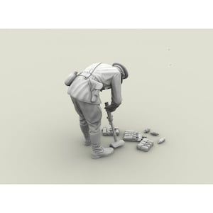 Könnte beinhalten: Eine monochrome, graustufige Miniaturfigur eines Soldaten in Uniform mit Mütze und Stiefeln, der sich nach vorne beugt, um eine mörserartige Waffe zu bedienen. Mehrere kleine Munitionsbehälter und Taschen liegen verstreut auf dem Boden.