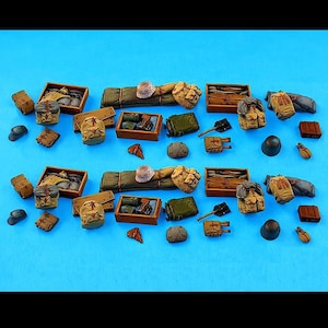 May include: A collection of miniature military equipment including brown wooden crates, tan backpacks, rolled sleeping mats, dark green metal canisters, small shovels, helmets, and various pouches arranged in two identical rows on a blue background.