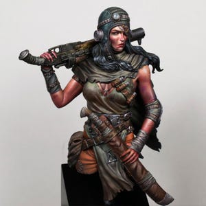 May include: A hand-painted resin bust of a post-apocalyptic character wearing a dark aviator cap with goggles, a tattered olive green top, and a brown leather holster. The figure carries a large rifle over one shoulder and a triangular pendant.