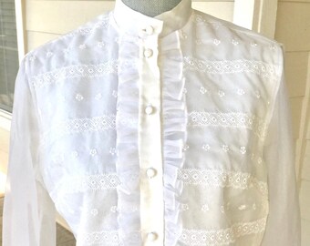 May include: White long-sleeved blouse with a high collar and button-up front. The blouse features delicate lace trim and a ruffled design.