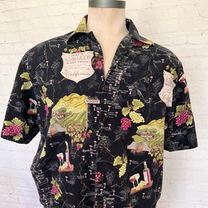 May include: A black short-sleeved button-up shirt with a map of the Napa Valley Wine Trail printed on it. The map features the names of wineries, vineyards, and towns. The shirt also has images of grapes, wine glasses, and wine bottles.