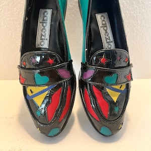 May include: A pair of black leather loafers with a teal blue heel. The loafers have a colorful design with red, yellow, blue, and purple accents. The loafers are decorated with stars and hearts. The loafers are labeled "Calpezio" on the inside.