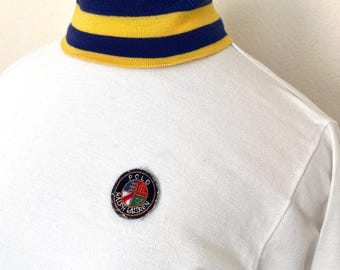 May include: White long-sleeve shirt with a yellow and navy blue striped turtleneck collar. A circular patch with the text "POLO RALPH LAUREN" is sewn on the chest. The shirt is displayed on a mannequin.