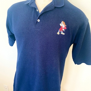 May include: A navy blue polo shirt with a cartoon character embroidered on the right side of the chest. The character is red, white, and blue and appears to be a dwarf with a beard and a hat.