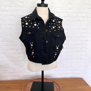 May include: A black denim vest with gold and clear rhinestones. The vest has a button closure and a classic denim style.