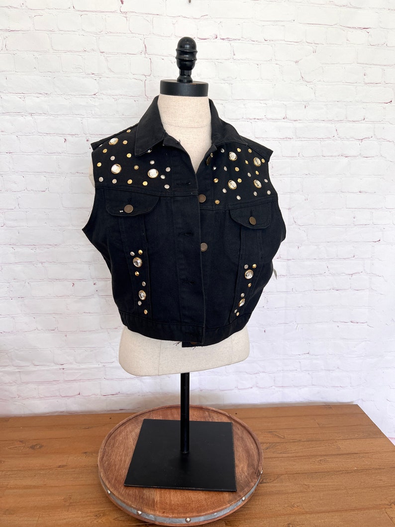 May include: A black denim vest with gold and clear rhinestones. The vest has a button closure and a classic denim style.