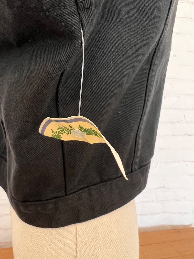 May include: Black denim jacket with a small tag attached to the bottom. The tag is cream colored with green leaves and blue stripes.