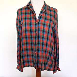 May include: A long-sleeved button-down shirt featuring a plaid pattern in shades of red, black, and blue. The shirt has a classic collar, a front button placket with small orange buttons, and long sleeves with buttoned cuffs, displayed on a mannequin.
