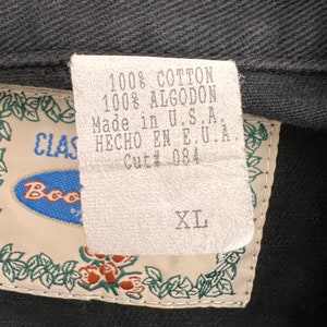 May include: A white tag with black text that reads "100% Cotton 100% Algodon Made in U.S.A. HECHO EN E.U.A. Cut# 084" and a white tag with black text that reads "XL". The tags are attached to a black garment with a green floral border.