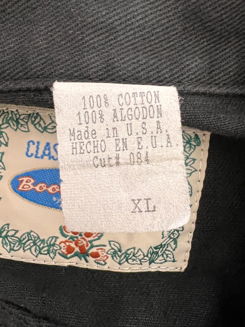 May include: A white tag with black text that reads "100% Cotton 100% Algodon Made in U.S.A. HECHO EN E.U.A. Cut# 084" and a white tag with black text that reads "XL". The tags are attached to a black garment with a green floral border.