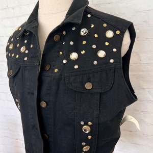 May include: A black denim vest with a cropped hem and sleeveless design. The vest is adorned with a variety of rhinestones and gold-toned buttons.