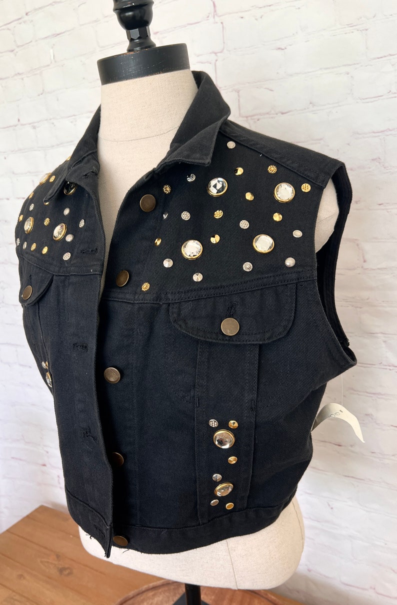 May include: A black denim vest with a cropped hem and sleeveless design. The vest is adorned with a variety of rhinestones and gold-toned buttons.