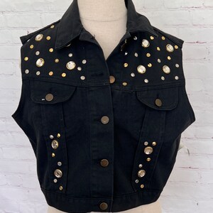 May include: A black denim vest with a button-up front and sleeveless design. The vest is decorated with gold and silver rhinestones.