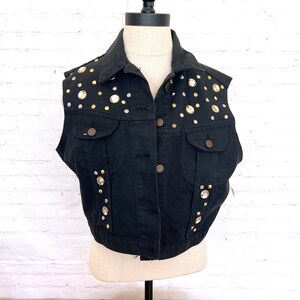 May include: A black denim vest with gold and clear rhinestone embellishments. The vest has a button closure and two pockets.