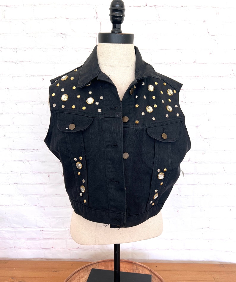 May include: A black denim vest with gold and clear rhinestone embellishments. The vest has a button closure and two pockets.
