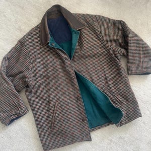 May include: A long-sleeve jacket featuring a brown, black, and red houndstooth pattern. The jacket has a dark brown leather collar, a button-front closure, and a solid dark teal interior lining with a quilted navy blue section at the upper back.