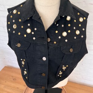 May include: A black denim vest with a cropped fit and a collar. The vest is decorated with silver and gold-toned rhinestones.