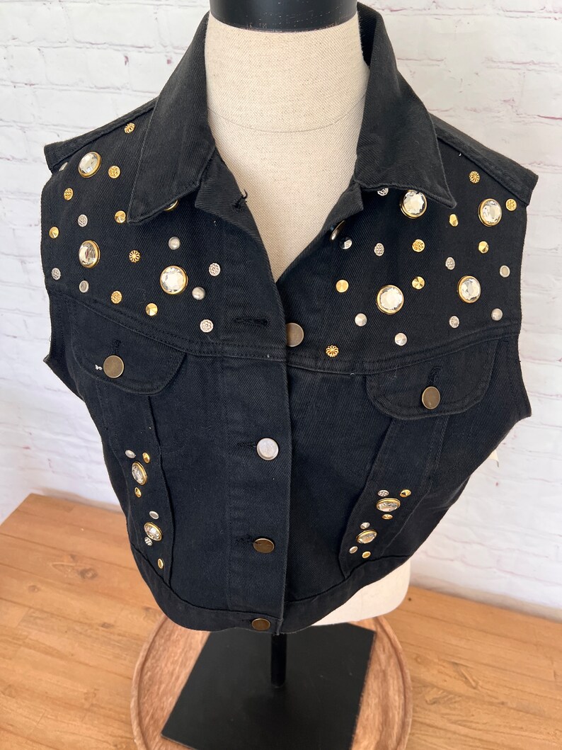 May include: A black denim vest with a cropped fit and a collar. The vest is decorated with silver and gold-toned rhinestones.