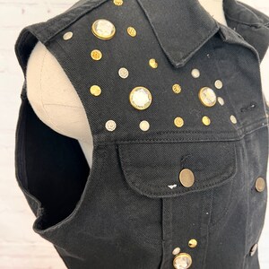May include: A black denim vest with a variety of rhinestones and gold-toned accents. The vest has a collar and a front pocket.