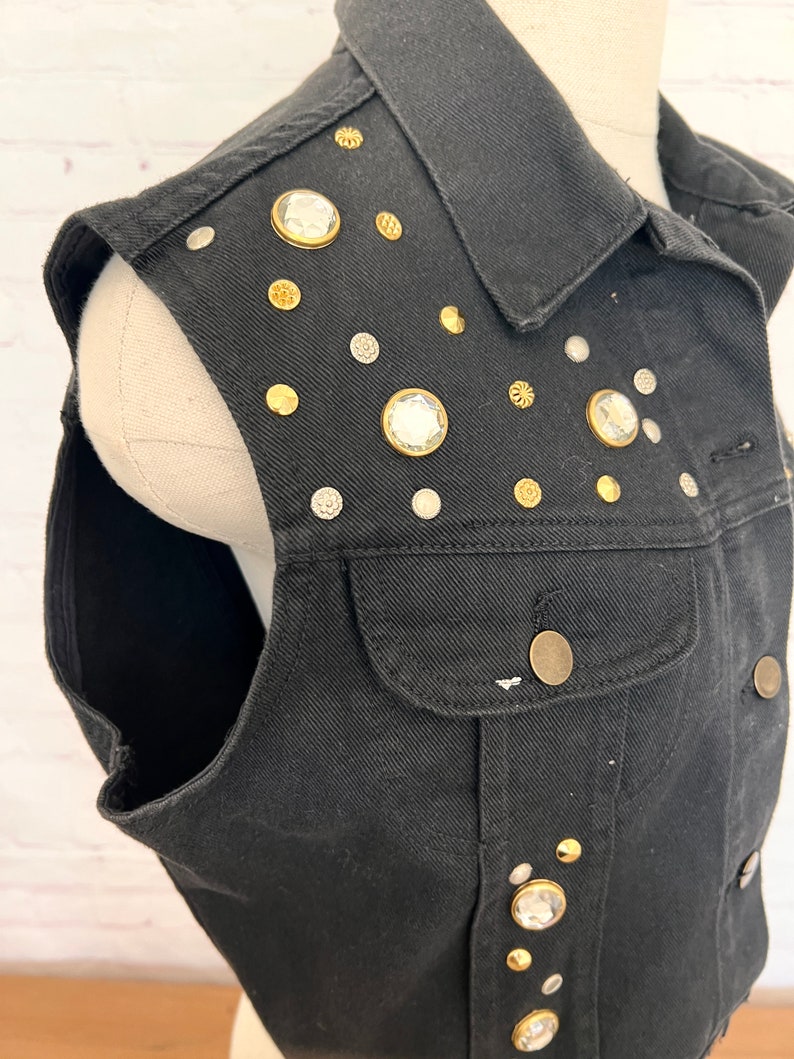 May include: A black denim vest with a variety of rhinestones and gold-toned accents. The vest has a collar and a front pocket.
