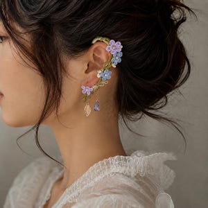May include: An ear cuff featuring green embroidered vines, light blue and purple fabric flowers with small white beads, a dangling gold-toned metal leaf charm, and a single translucent purple teardrop-shaped crystal bead accent.