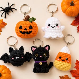 May include: Five crochet keychains featuring Halloween themes: an orange jack-o'-lantern, a white ghost, a black bat with white fangs, a black cat with a purple collar and gold bell, and a yellow, orange, and white candy corn, all with metal rings.