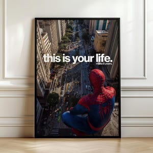 Puede incluir: Un póster vertical enmarcado que muestra a un personaje con traje rojo y azul sentado en una cornisa sobre una calle urbana. El texto "this is your life. make it yours." está impreso en blanco sobre la escena con edificios y tráfico.