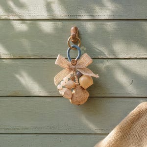 May include: A decorative hanging ornament featuring a blue wrapped ring, a tan burlap bow, and a cluster of assorted wooden and smooth beads in shades of cream, brown, and gray, suspended from a small wooden hook on a light green wooden wall.