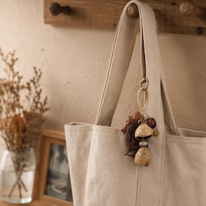 Puede incluir: Un bolso tote de lona beige con un colgante que presenta elementos naturales, incluyendo una vaina de semilla curva de color marrón oscuro, dos cáscaras de nuez texturizadas de color marrón claro, una cuenta gris lisa y una cuenta verde.