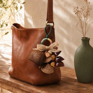 May include: A brown leather hobo bag with a decorative charm attached to the strap. The charm features a green and yellow wrapped ring, a beige burlap bow, a textured brown coconut shell, and several small dark brown wooden bell-shaped pieces.