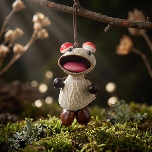 Può includere: Un ornamento sospeso realizzato con materiali naturali, caratterizzato da una testa in baccello testurizzato di colore chiaro, una bocca spalancata, una lingua rosa e due piccoli cappelli di fungo rossi come orecchie.