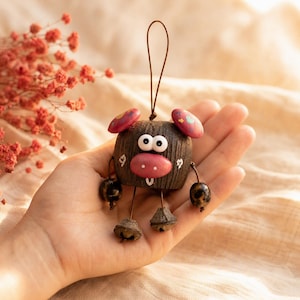 May include: A small, dark brown, textured wooden ornament shaped like a pig with a pink snout, two large white eyes, and pink ears decorated with small colourful shapes. It features dangling limbs with dark beads and metal bell feet on a cord.