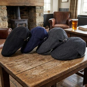 May include: Four flat caps arranged in a row on a wooden surface. The caps are made of a textured fabric in black, navy blue, and two shades of gray. Each cap features a side adjustment strap with a small metal buckle for a secure and adjustable fit.