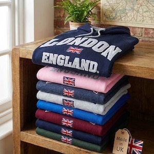 Puede incluir: Una pila de ocho sudaderas con capucha de manga larga en varios colores, incluyendo azul marino, rosa, gris, azul, granate y verde. Cada sudadera presenta una bandera Union Jack bordada y el texto LONDON ENGLAND en la prenda superior.