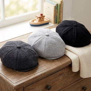 May include: Three newsboy caps in different colors and textures. One cap is dark gray with a herringbone pattern, one is solid light gray, and one is solid black. Each cap features a rounded crown with a central button and a short front brim.