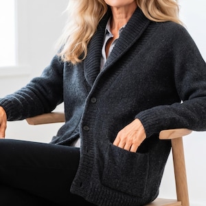May include: A charcoal gray knit cardigan featuring a wide shawl collar, long sleeves with ribbed cuffs, and a button-down front closure. The garment includes two large patch pockets on the front and is worn over a light-colored collared shirt.