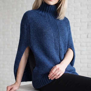 Peut inclure: Un poncho en tricot bleu foncé avec un col roulé haut, présentant une texture mouchetée sur tout le tissu. Le vêtement possède de larges ouvertures pour les bras sur les côtés et un ourlet côtelé en bas, offrant une coupe structurée et ample.
