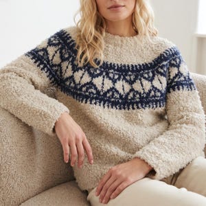 May include: A long-sleeved, cream-colored sweater with a textured, fuzzy fleece-like material. The garment features a dark navy blue geometric pattern across the chest and shoulders, extending down the sleeves in a circular yoke design.