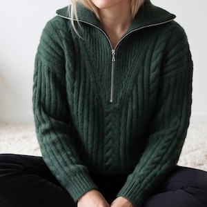 May include: A dark forest green quarter-zip jumper featuring a ribbed knit texture and vertical cable knit detailing. The garment includes a high collar with a silver-toned metal zip closure and long sleeves with ribbed cuffs at the wrists.