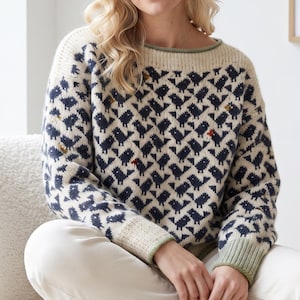 May include: A cream-colored long-sleeve sweater featuring a repeating dark blue bird pattern. The knit garment has a wide neckline, ribbed cuffs in a muted sage green, and small, scattered orange and yellow accents throughout the bird design.