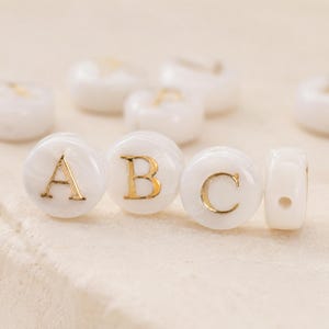 May include: A collection of round, white, marbled beads featuring embossed gold-coloured letters. Three beads in the foreground clearly display the letters A, B, and C in a serif font, while other beads are scattered in the background.