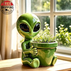 May include: A glossy green ceramic planter shaped like an alien figure with large black eyes, holding a round pot containing small green plants. The alien has a smooth, reflective surface and is seated in a relaxed position with its arms wrapped around the pot.